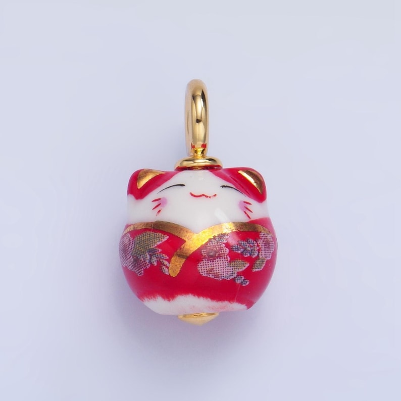 May include: A red and white ceramic charm in the shape of a smiling cat wearing a red and gold patterned kimono. The charm has a gold loop for hanging.