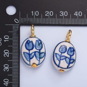 May include: Two blue and white ceramic pendants with gold accents. Each pendant features a design of two blue tulips with green stems and leaves. The pendants are oval shaped and have a gold loop at the top for hanging.