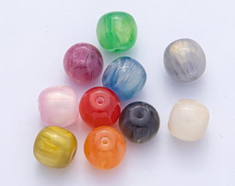 Puede incluir: Una colección de diez cuentas redondeadas de colores, incluyendo verde, rosa, rojo, azul, naranja, amarillo, gris y negro. Las cuentas tienen un aspecto veteado o arremolinado y están dispersas sobre una superficie blanca.
