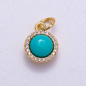 May include: A gold-colored pendant with a round, bright turquoise center stone. The stone is surrounded by a halo of small, clear crystals. The pendant has a small loop at the top for attaching to a chain or bracelet. Jewelry.