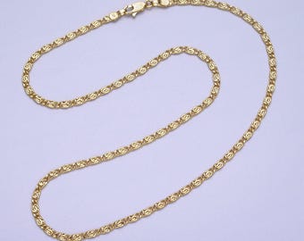 May include: A gold-colored necklace with a delicate, repeating pattern. The chain is made of small, interconnected links, creating a textured appearance. The necklace has a lobster clasp closure.