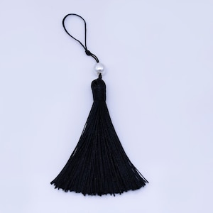 Könnte beinhalten: Eine schwarze Quaste mit einer Schlaufe und einer silbernen Perle. Die Quaste besteht aus vielen schwarzen Fäden, und die Perle ist rund und glänzend. Die Quaste befindet sich vor einem weißen Hintergrund.