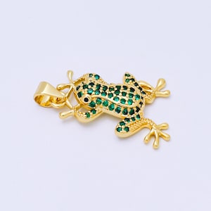 May include: A gold-colored frog pendant with green gemstone accents. The frog is detailed with small green stones covering its back and a single black stone for the eye. The pendant has a loop for attaching to a chain.