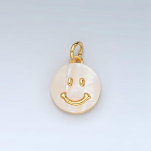 May include: A gold-colored pendant with a smiley face design. The pendant features a round, off-white face with a gold-colored, smiling mouth and two eyes. The pendant has a gold-colored loop at the top for attaching to a chain.