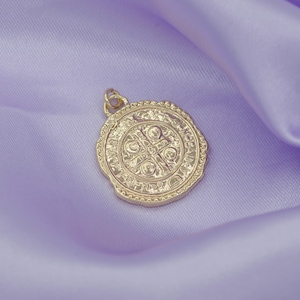 May include: A gold-toned pendant with a detailed design featuring a cross and other symbols. The pendant is on a purple fabric background.