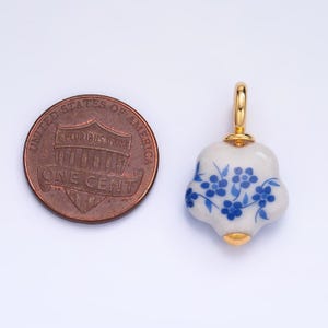 Op de afbeelding: Een kleine, witte porseleinen hanger met een blauw bloemenmotief. De hanger heeft een gouden lus aan de bovenkant en is ongeveer zo groot als een Nederlandse cent.