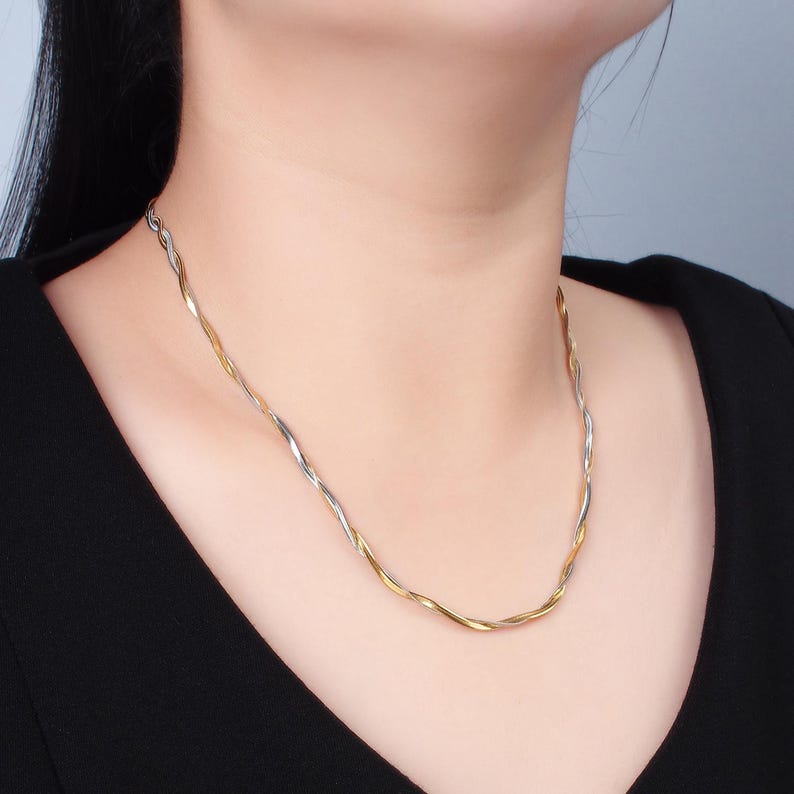 May include: A silver and gold twisted chain necklace. The necklace is worn by a person with dark hair and a black shirt.