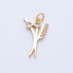 May include: A gold-colored pendant featuring a pair of scissors and a comb, with two clear, round gemstones set into the scissors. The pendant has a loop at the top for attaching to a chain or bracelet. The design is a charm for hairstylists.