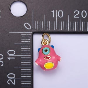 May include: A pink enamel monster charm with a blue eye, yellow mouth, and yellow spot. The charm is attached to a gold jump ring.