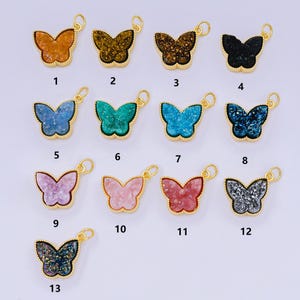 Puede incluir: Una colección de colgantes con forma de mariposa en varios colores, incluyendo naranja, marrón, negro, azul, verde, rosa y rojo. Cada colgante tiene una anilla dorada para sujetarlo a una cadena. Los colgantes probablemente son de piedra o resina.