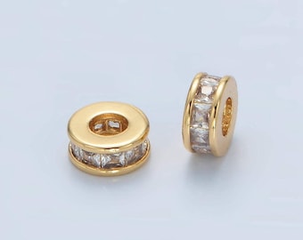 May include: Two gold-colored, cylindrical jewelry beads. Each bead features a ring of clear, square-cut stones set between two gold bands. The beads have a hole through the center, and are displayed against a light background.