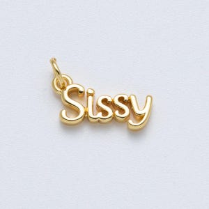 May include: A gold-colored charm pendant with the word "Sissy" in a cursive font. The charm has a small loop at the top for attaching to a necklace or bracelet. The background is a plain white surface.
