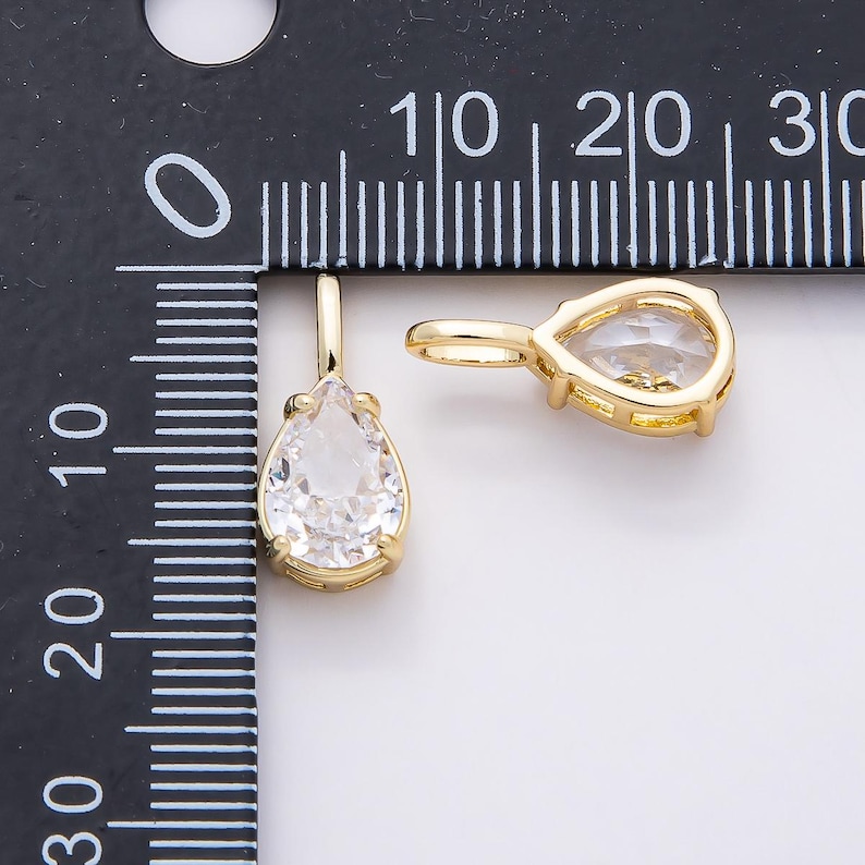 Op de afbeelding: Twee goudkleurige, druppelvormige hangers met heldere, facetgeslepen stenen. De hangers worden getoond tegen een witte achtergrond, met een liniaal op de achtergrond voor de schaal. Dit zijn waarschijnlijk sieraden.