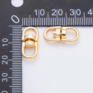 May include: Two gold-colored swivel clasps. Each clasp has a D-ring on either end, connected by a cylindrical center piece. The clasps are shown against a white background with a black ruler, providing scale. These clasps are likely used for jewelry or bag making.
