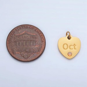 May include: Gold heart-shaped charm with the word "Oct" engraved on it. A small, pale pink gemstone is set in the center of the charm. The charm is next to a US penny for size comparison.