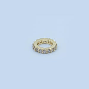 Könnte beinhalten: Ein goldfarbener Ring mit einer durchgehenden Reihe klarer, quadratisch geschliffener Edelsteine. Der Ring ist vor einem hellblauen Hintergrund platziert, der sein kreisförmiges Design und die funkelnden Steine hervorhebt. Das Ringband ist ebenfalls goldfarben.