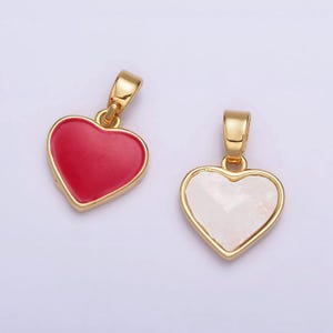 Op de afbeelding: Twee gouden hartvormige hangers, een met een rode emaille afwerking en de andere met een witte, iriserende afwerking.