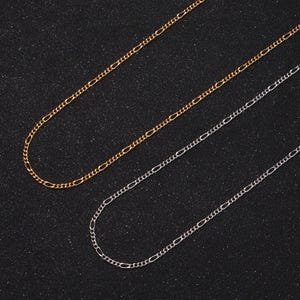 May include: Two gold and one silver chain necklaces on a black background. The chains are all the same style, with a simple, flat link design.