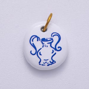 May include: White, oval pendant featuring a blue vase illustration. A gold-coloured loop is attached at the top for suspension. The vase design is a simple, stylised drawing.