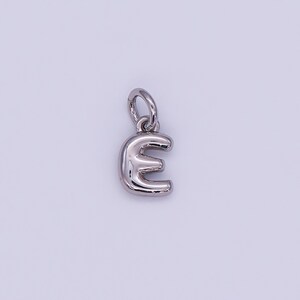 May include: A silver-colored pendant in the shape of the letter "E". The pendant has a rounded, three-dimensional design with a small loop at the top for attaching to a chain or bracelet. The background is a solid, light color.