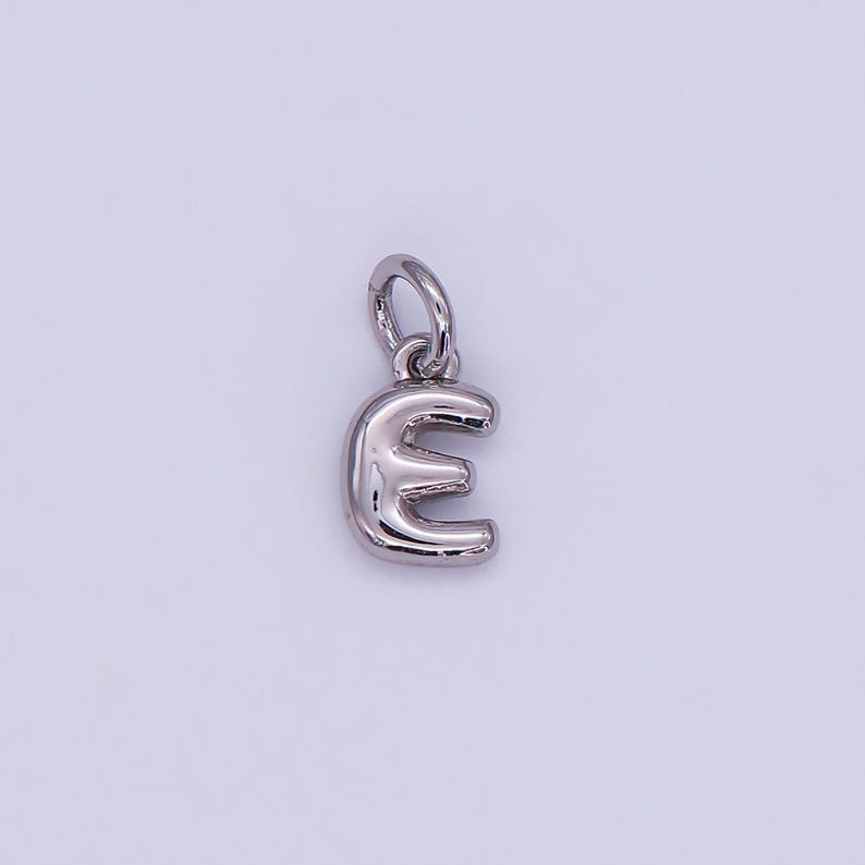 May include: A silver-colored pendant in the shape of the letter "E". The pendant has a rounded, three-dimensional design with a small loop at the top for attaching to a chain or bracelet. The background is a solid, light color.