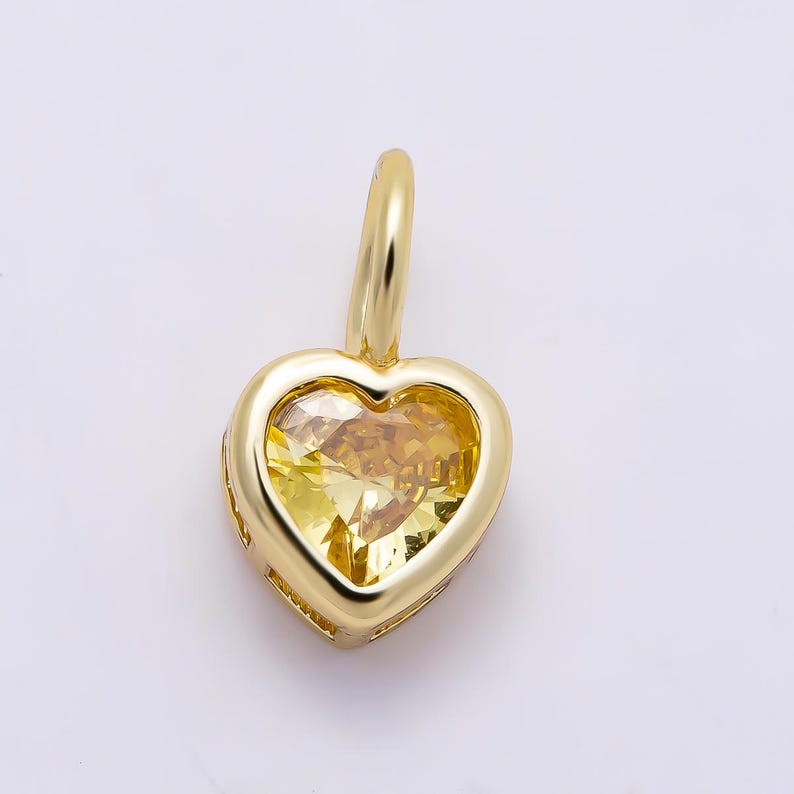 May include: A gold pendant featuring a heart-shaped yellow gemstone set within a polished gold frame. The pendant includes a small, rounded gold bail at the top for attachment, showcasing a faceted cut that reflects light across the yellow stone.