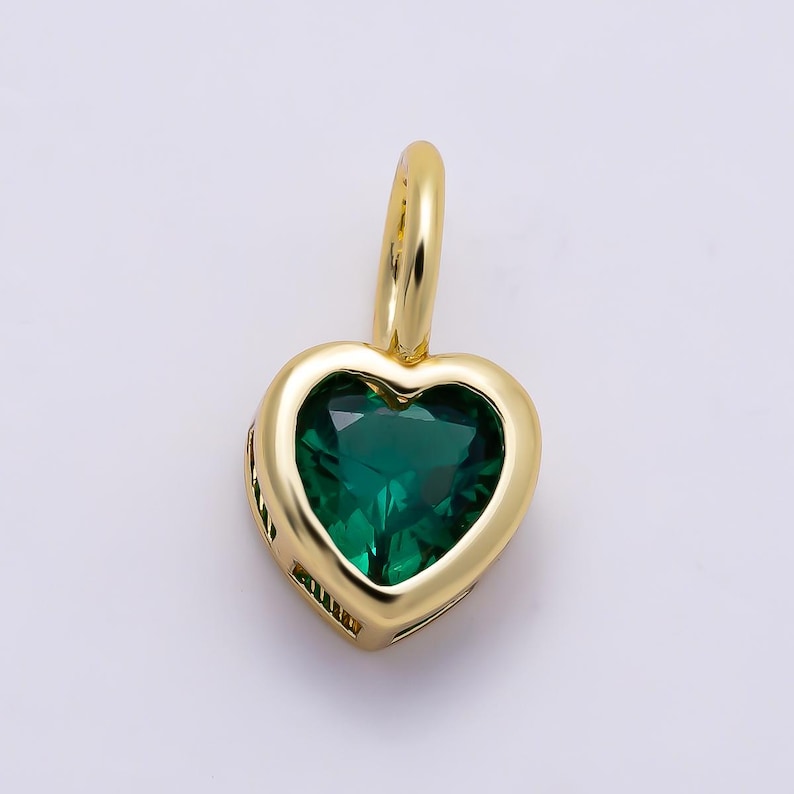 May include: A heart-shaped pendant featuring a faceted deep green gemstone set within a polished gold-toned metal frame. The pendant includes a small, smooth gold-toned bail at the top for attachment to a chain, showcasing a clean, reflective finish.