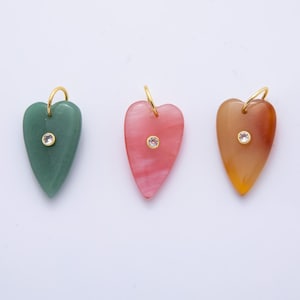 Puede incluir: Tres colgantes en forma de corazón en verde, rosa y naranja, cada uno con un lazo dorado y una pequeña gema transparente. Los colgantes son lisos y pulidos, con una ligera variación de color y textura.