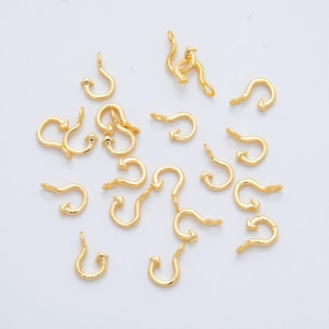 May include: A collection of small, gold-colored jewelry findings, each with a curved hook shape and an arrow-like detail. The findings are scattered across a white surface, suggesting a craft or jewelry-making theme.