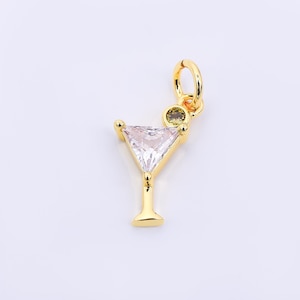 May include: A gold-colored charm in the shape of a cocktail glass. The glass features a clear, triangular-cut gemstone and a small, round, olive-colored stone. The charm has a loop for attaching to a necklace or bracelet.