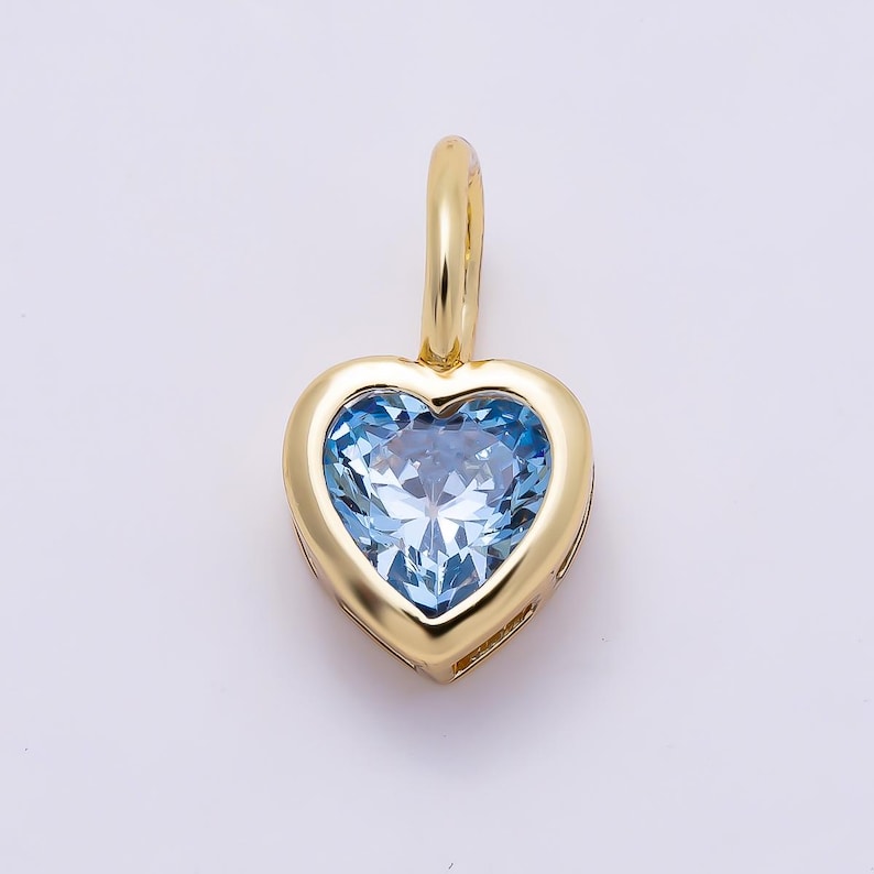 May include: A heart-shaped pendant featuring a faceted light blue gemstone set in a polished yellow gold frame. The pendant includes a small, smooth yellow gold bail at the top, designed for attachment to a necklace chain or cord.