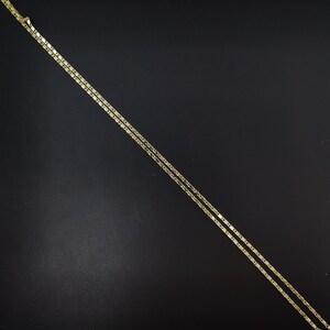 May include: A delicate gold-colored necklace with a flat, rectangular link chain design. The necklace is displayed against a dark background, highlighting its subtle shine. The chain is thin and appears to be made of metal.