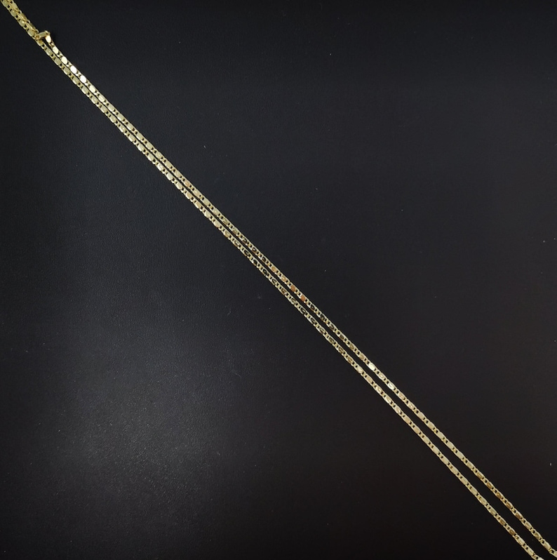May include: A delicate gold-colored necklace with a flat, rectangular link chain design. The necklace is displayed against a dark background, highlighting its subtle shine. The chain is thin and appears to be made of metal.