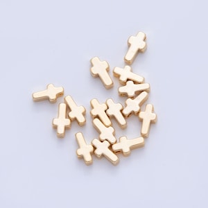 May include: A collection of small, gold-colored cross-shaped beads. The beads are scattered across a light-colored surface, each with a simple, rounded design. These beads are suitable for jewelry making or craft projects.
