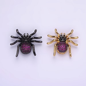 Puede incluir: Dos abalorios de araña con acabados en negro y dorado. Ambos abalorios tienen un cuerpo de pedrería morada y ojos de pedrería negra.