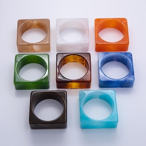 Può includere: Una collezione di braccialetti quadrati e spessi in vari colori. I braccialetti sono realizzati in un materiale traslucido e includono sfumature di marrone, bianco, arancione, verde, ambra, blu, marrone scuro e turchese. Sono disposti su una superficie bianca.