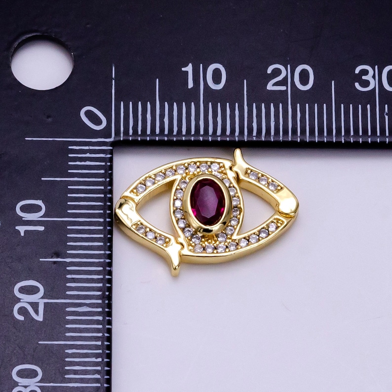 May include: Gold-tone eye charm with a red gemstone center and clear rhinestones around the eye and the perimeter. The charm is approximately 2 cm long.