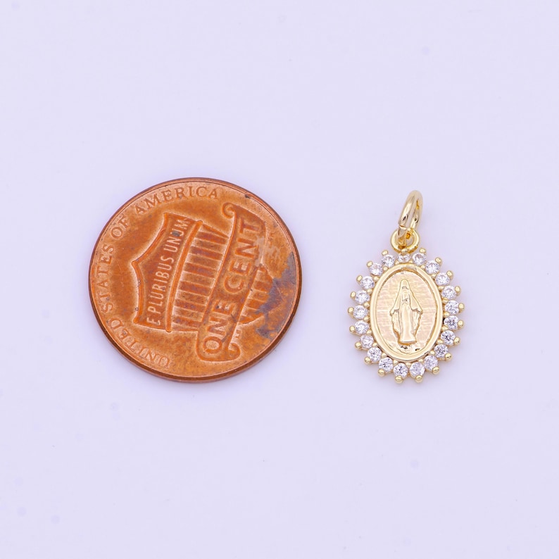 May include: Gold-tone pendant featuring a depiction of the Virgin Mary with a halo of clear cubic zirconia stones. The pendant is shown next to a US penny for size comparison.