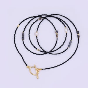May include: A long, layered necklace featuring black beads, gold accents, and natural stone and pearl embellishments. The necklace has a gold toggle clasp. The design incorporates rectangular and square stones.
