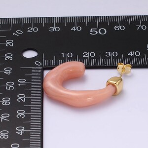 May include: A single peach-colored, curved hoop earring with a gold-colored post and square accent. The earring is set against a white background with a black ruler for scale.