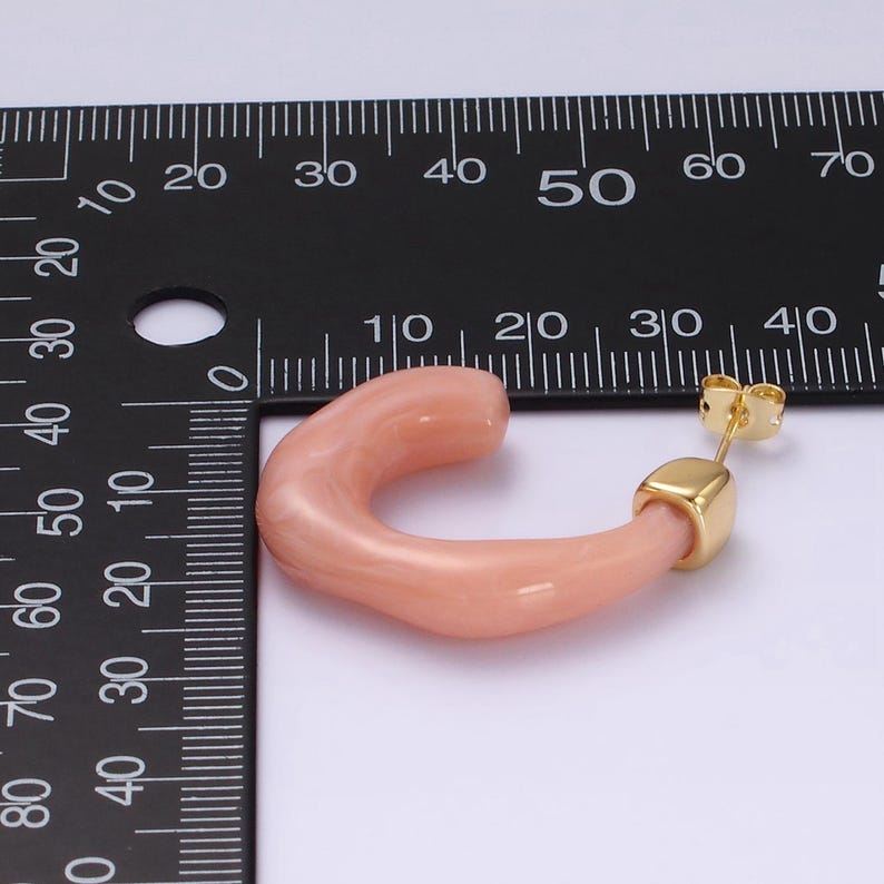 May include: A single peach-colored, curved hoop earring with a gold-colored post and square accent. The earring is set against a white background with a black ruler for scale.