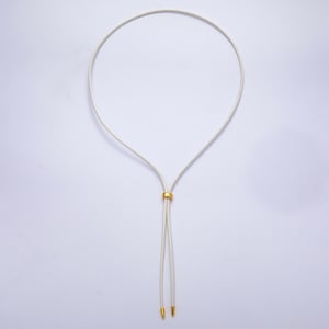 K&ouml;nnte beinhalten: Eine cremefarbene Halskette mit einem goldfarbenen, kreisf&ouml;rmigen Akzent. Die Halskette hat ein schlichtes, elegantes Design mit zwei herabh&auml;ngenden Enden, die jeweils mit einem goldfarbenen Detail versehen sind. Die Halskette wird vor wei&szlig;em Hintergrund pr&auml;sentiert.