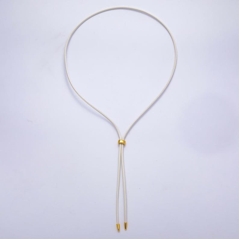 K&ouml;nnte beinhalten: Eine cremefarbene Halskette mit einem goldfarbenen, kreisf&ouml;rmigen Akzent. Die Halskette hat ein schlichtes, elegantes Design mit zwei herabh&auml;ngenden Enden, die jeweils mit einem goldfarbenen Detail versehen sind. Die Halskette wird vor wei&szlig;em Hintergrund pr&auml;sentiert.