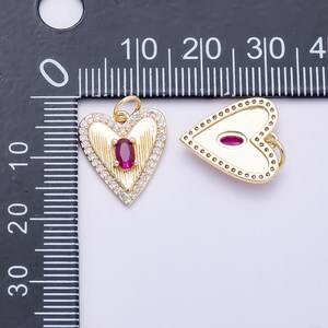 May include: Two gold heart-shaped pendants with a jump ring. The left pendant features a central oval red stone, textured lines, and a border of small clear stones. The right pendant has a smooth surface with a central oval red stone and a dotted border.