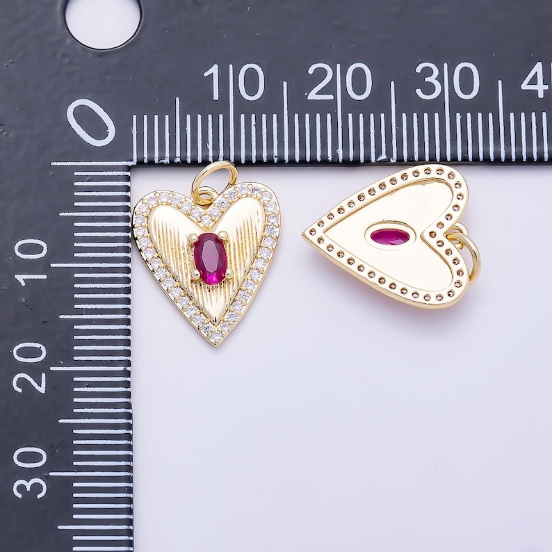 May include: Two gold heart-shaped pendants with a jump ring. The left pendant features a central oval red stone, textured lines, and a border of small clear stones. The right pendant has a smooth surface with a central oval red stone and a dotted border.