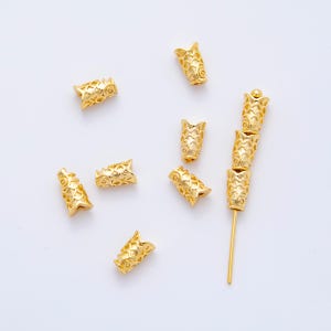 May include: Gold-colored metal beads with a fish-scale pattern. The beads are cylindrical with a flared top and bottom. Some beads are scattered, while others are strung on a gold-colored metal rod. The beads are likely for jewelry making.