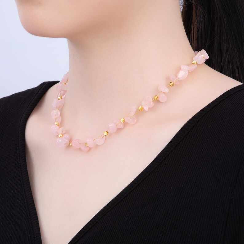 May include: A delicate necklace featuring rose quartz chips and small gold-colored beads. The pink stones are strung together, creating a unique and elegant accessory. The necklace is shown against a black top.