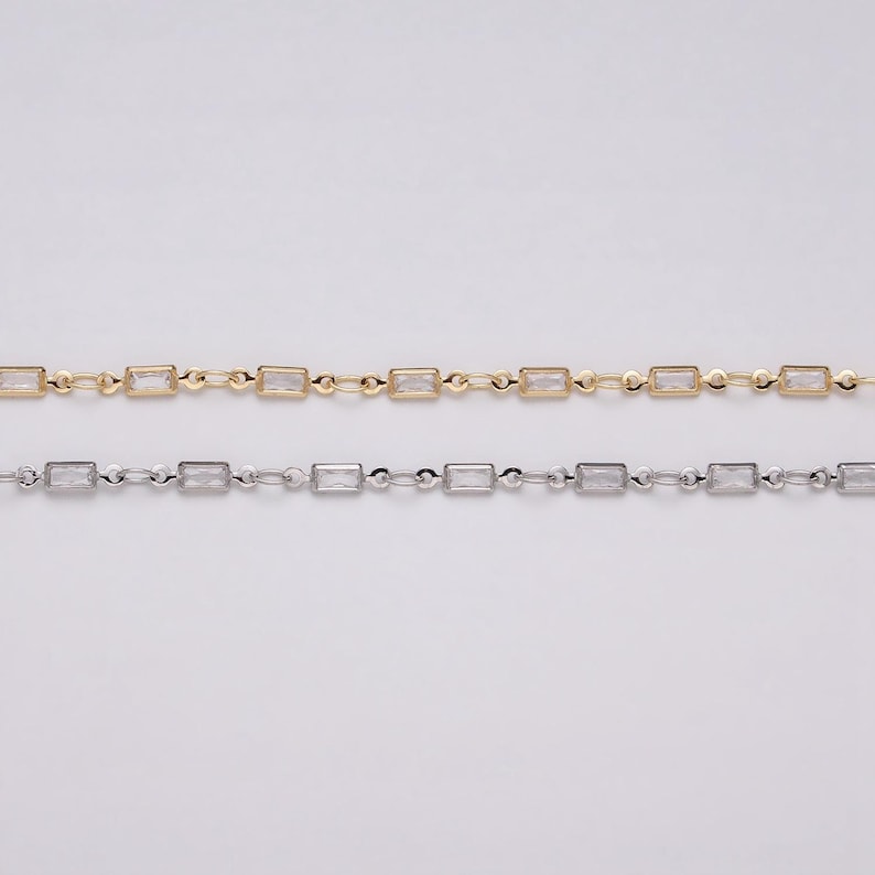 May include: Two delicate necklaces, one gold-toned and one silver-toned, each featuring rectangular, clear gemstones. The gemstones are evenly spaced along the chain, creating a refined and elegant look. The chains are displayed against a neutral background.