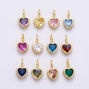 May include: A collection of twelve heart-shaped pendants, each featuring a faceted gemstone in a different color set within a gold-toned metal frame. Each pendant includes a small gold-toned jump ring at the top for attachment to a chain or cord.