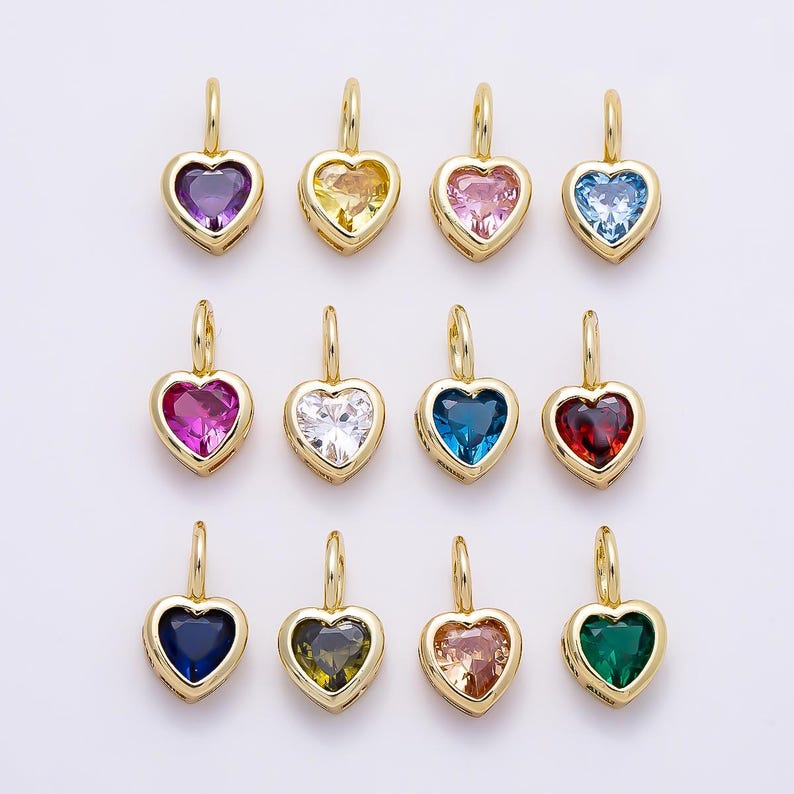 May include: A collection of twelve heart-shaped pendants, each featuring a faceted gemstone in a different color set within a gold-toned metal frame. Each pendant includes a small gold-toned jump ring at the top for attachment to a chain or cord.